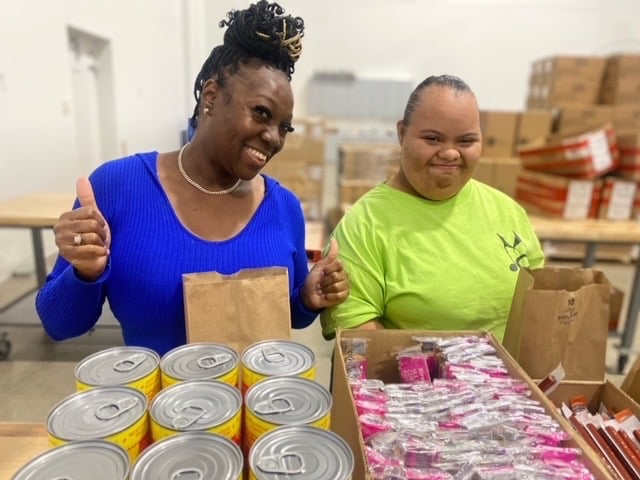 Two people smiling behind cans and packages of food in the volunteer packing area at Freestore Foodbank.